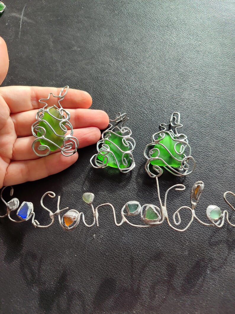 May include: Three green glass Christmas tree ornaments wrapped in silver wire. The ornaments are hanging from a silver wire that spells out the word "Sea Glass".