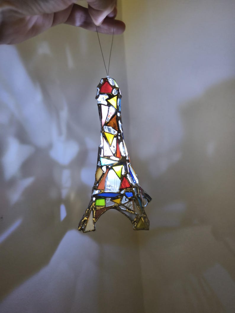 May include: A stained glass Eiffel Tower ornament in a variety of colours. The miniature tower is made from pieces of red, yellow, blue, and white glass. It is suspended by a thin wire.