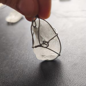May include: A clear, irregularly shaped pendant wrapped in dark wire. The pendant is held by a hand against a dark background. The wire wraps around the stone, creating a cage-like design. The pendant appears to be made of a translucent material.