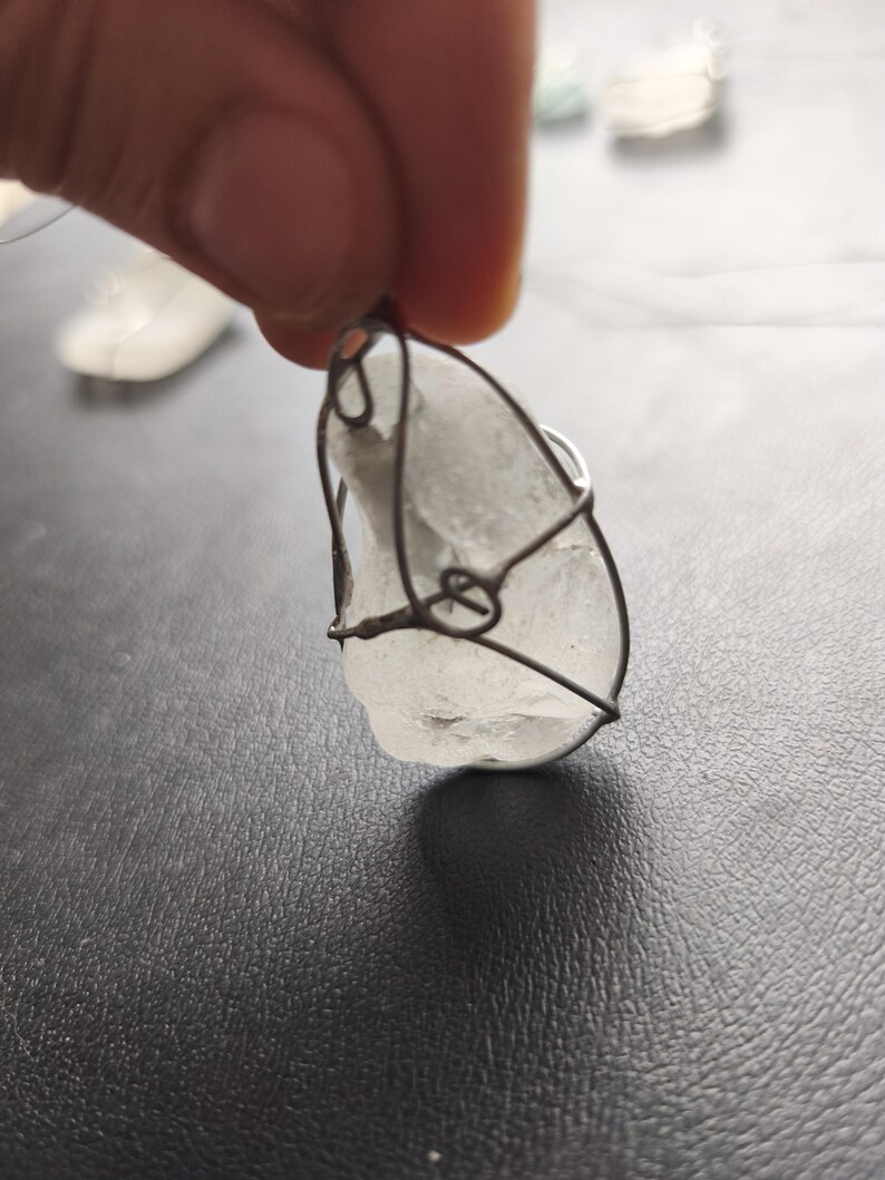 May include: A clear, irregularly shaped pendant wrapped in dark wire. The pendant is held by a hand against a dark background. The wire wraps around the stone, creating a cage-like design. The pendant appears to be made of a translucent material.