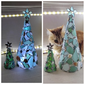 Pode incluir: Duas &aacute;rvores de Natal decorativas feitas de vidro do mar. A &aacute;rvore maior &eacute; verde-azulado e azul, com uma estrela no topo. A &aacute;rvore menor &eacute; verde. Ambas as &aacute;rvores t&ecirc;m formato de cone e s&atilde;o iluminadas com luzes internas.
