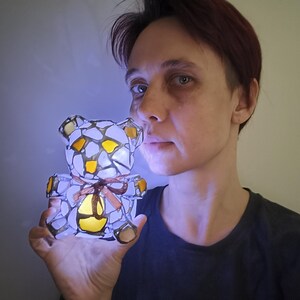 May include: A glowing stained glass teddy bear lamp with a brown ribbon. The bear is made of white, yellow, and brown glass pieces. The lamp is lit from within, creating a soft, warm glow. The bear is held up in front of a person.