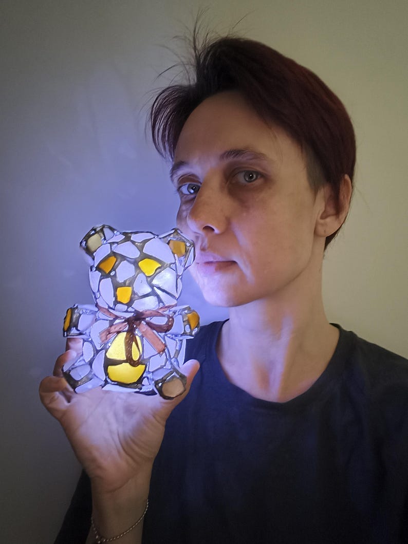 May include: A glowing stained glass teddy bear lamp with a brown ribbon. The bear is made of white, yellow, and brown glass pieces. The lamp is lit from within, creating a soft, warm glow. The bear is held up in front of a person.