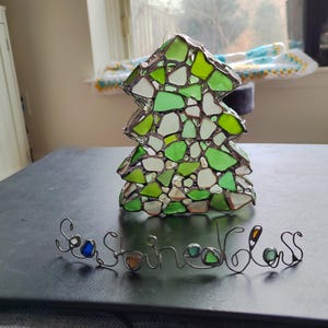 K&ouml;nnte beinhalten: Ein gr&uuml;ner Weihnachtsbaum-Anh&auml;nger aus Glas, der aus kleinen St&uuml;cken Meerglas besteht. Der Anh&auml;nger liegt auf einer schwarzen Oberfl&auml;che mit einer Drahtschrift, die "Sea Glass" lautet.