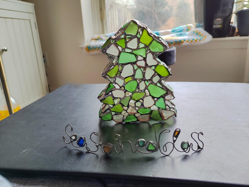 K&ouml;nnte beinhalten: Ein gr&uuml;ner Weihnachtsbaum-Anh&auml;nger aus Glas, der aus kleinen St&uuml;cken Meerglas besteht. Der Anh&auml;nger liegt auf einer schwarzen Oberfl&auml;che mit einer Drahtschrift, die "Sea Glass" lautet.