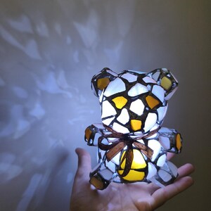 May include: A stained glass teddy bear lamp with a brown ribbon. The lamp is made of white, yellow, and brown glass pieces. The lamp is lit from within, casting a warm glow.