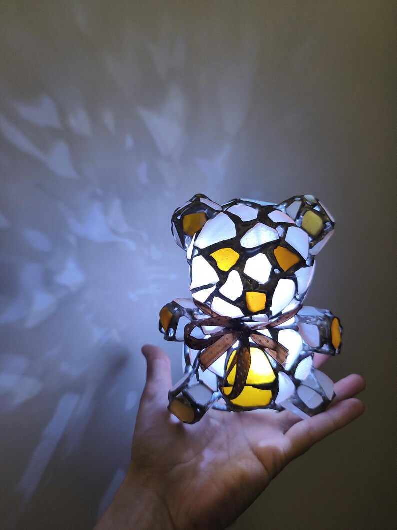 May include: A stained glass teddy bear lamp with a brown ribbon. The lamp is made of white, yellow, and brown glass pieces. The lamp is lit from within, casting a warm glow.