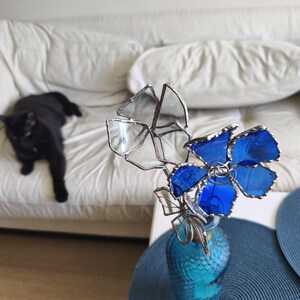 May include: A glass flower arrangement in a turquoise vase. The flowers are made of clear and blue glass with silver-coloured metal edging. A black cat is lounging on a white sofa in the background.