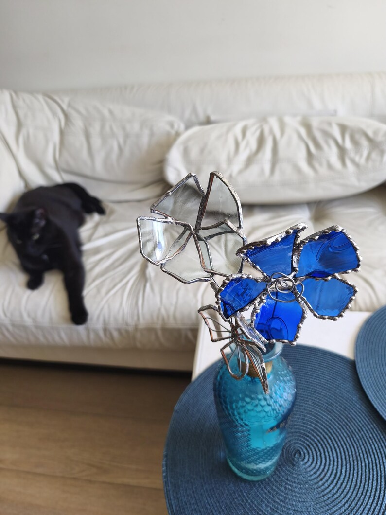 May include: A glass flower arrangement in a turquoise vase. The flowers are made of clear and blue glass with silver-coloured metal edging. A black cat is lounging on a white sofa in the background.