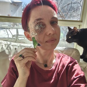 Puede incluir: Una persona con una camiseta burdeos, un collar de plata con una piedra verde y anillos con piedras verdes y rojas. Sostiene una pieza decorativa de alambre y vidrio para el ojo y un objeto de vidrio verde. Un gato negro est&aacute; cerca.
