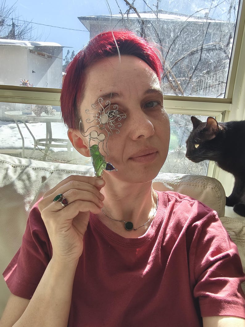Puede incluir: Una persona con una camiseta burdeos, un collar de plata con una piedra verde y anillos con piedras verdes y rojas. Sostiene una pieza decorativa de alambre y vidrio para el ojo y un objeto de vidrio verde. Un gato negro est&aacute; cerca.