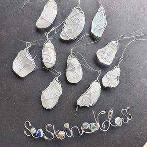 May include: A collection of sea glass pendants, each piece of frosted glass is wrapped in silver wire. The pendants vary in shape and size, with a wire loop for hanging. The words "Sea Stained Glass" are spelled out in wire.