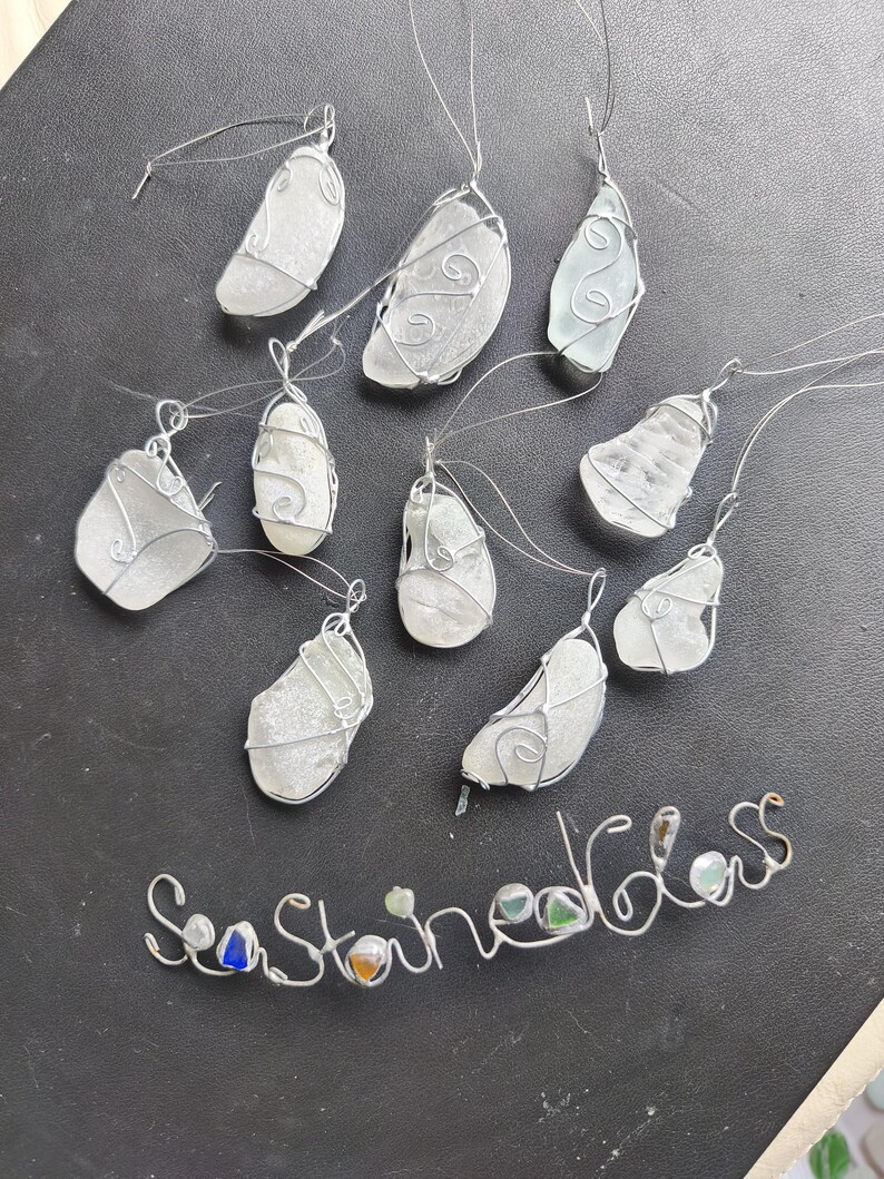 May include: A collection of sea glass pendants, each piece of frosted glass is wrapped in silver wire. The pendants vary in shape and size, with a wire loop for hanging. The words "Sea Stained Glass" are spelled out in wire.