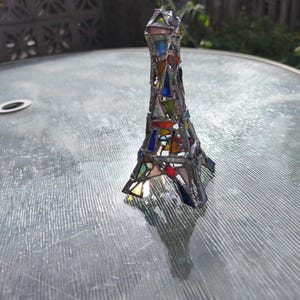 May include: A stained glass Eiffel Tower sculpture with a silver frame. The tower is made of colourful triangular glass pieces in shades of red, blue, green, and yellow. A thin wire extends from the top.