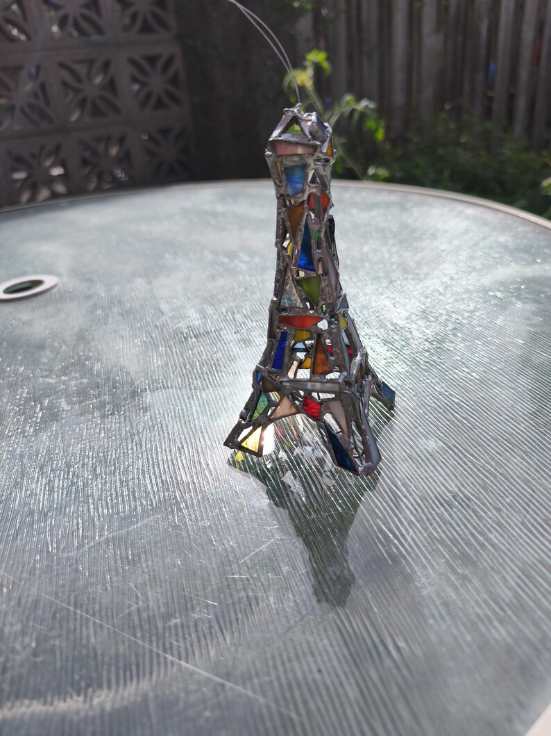 May include: A stained glass Eiffel Tower sculpture with a silver frame. The tower is made of colourful triangular glass pieces in shades of red, blue, green, and yellow. A thin wire extends from the top.