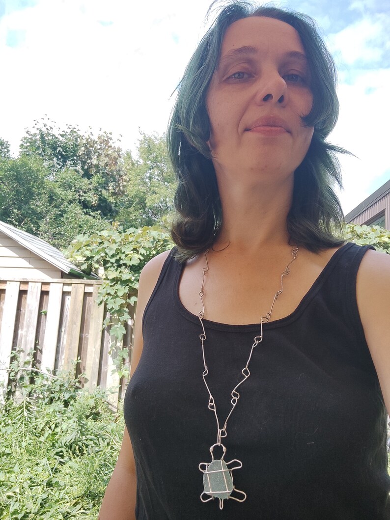 K&ouml;nnte beinhalten: Eine Frau mit blaugr&uuml;nem Haar tr&auml;gt ein schwarzes Tanktop und eine silberne Kettenkette mit einem gr&uuml;nen Steint&uuml;r-Anh&auml;nger.
