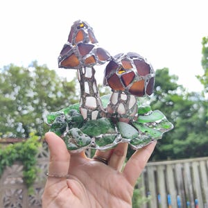 May include: A handcrafted stained glass mushroom sculpture. The mushrooms have brown and amber caps, white stems, and sit on a base of green glass pieces. The sculpture is held in a hand, with a blurred background of greenery.