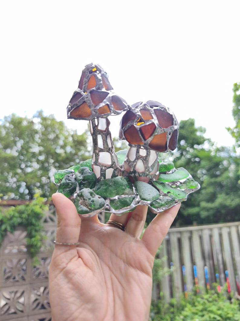 May include: A handcrafted stained glass mushroom sculpture. The mushrooms have brown and amber caps, white stems, and sit on a base of green glass pieces. The sculpture is held in a hand, with a blurred background of greenery.