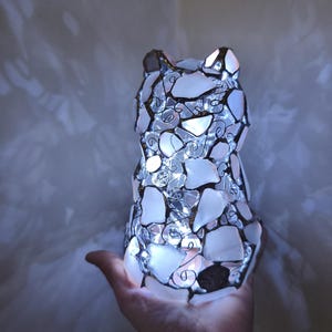 May include: A cat-shaped lamp made from white and clear glass pieces, with a wire frame and internal lights. The lamp is lit, producing a soft glow. The lamp is held in a hand.