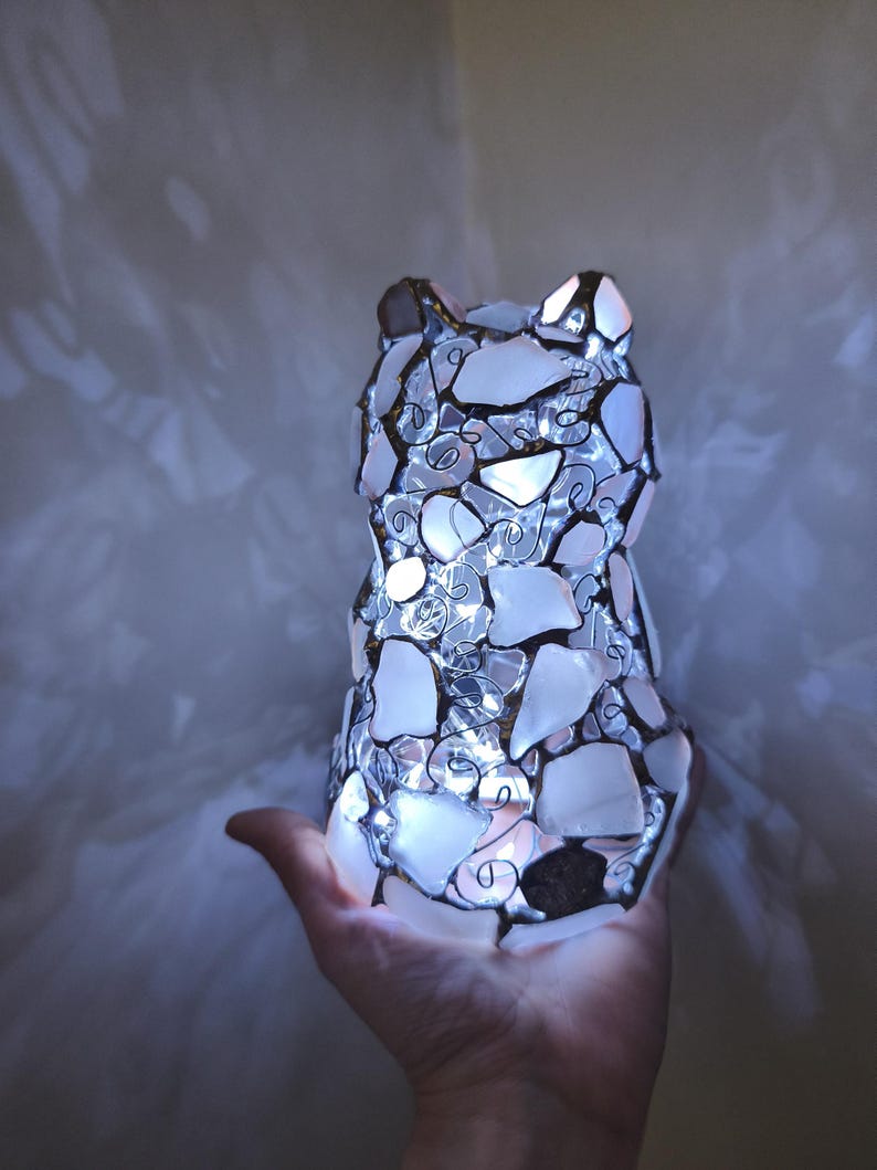 May include: A cat-shaped lamp made from white and clear glass pieces, with a wire frame and internal lights. The lamp is lit, producing a soft glow. The lamp is held in a hand.