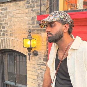 May include: A patterned cap with a colorful design is worn by a person. The person is wearing a black tank top and a cream-colored button-down shirt. A small brown bag hangs from the person's shoulder. The background includes a brick wall and a red door.