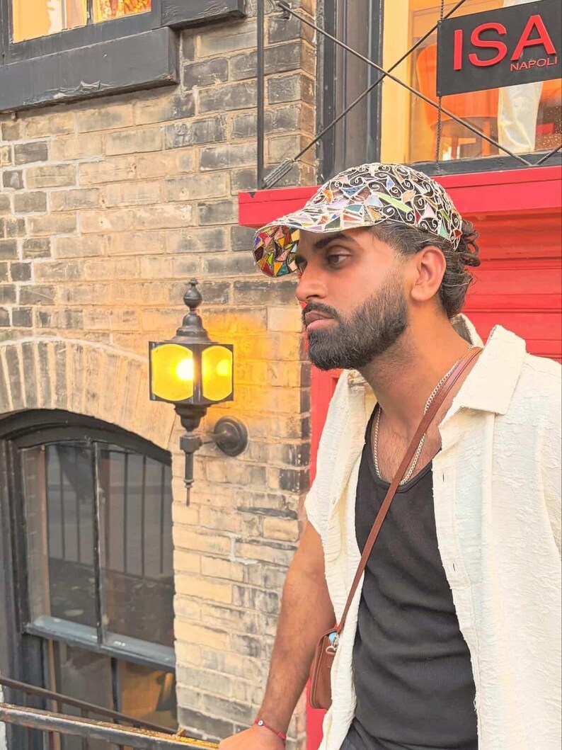 May include: A patterned cap with a colorful design is worn by a person. The person is wearing a black tank top and a cream-colored button-down shirt. A small brown bag hangs from the person's shoulder. The background includes a brick wall and a red door.