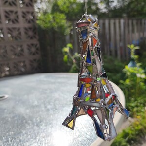 May include: A stained glass Eiffel Tower ornament, featuring a mosaic of coloured glass pieces. The glass includes red, blue, yellow, and green hues, set within a silver metal frame. The ornament hangs from a thin wire.