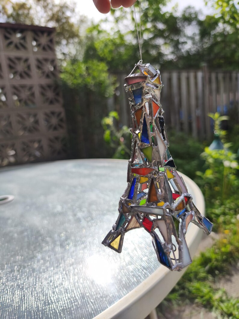 May include: A stained glass Eiffel Tower ornament, featuring a mosaic of coloured glass pieces. The glass includes red, blue, yellow, and green hues, set within a silver metal frame. The ornament hangs from a thin wire.
