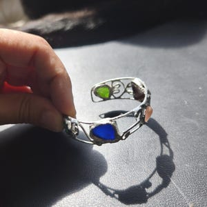 May include: Silver-toned cuff bracelet with colorful glass inserts. The bracelet features green, blue, and peach-colored glass pieces set within a decorative wire frame. The open design allows for easy wear and showcases the vibrant colors of the glass.