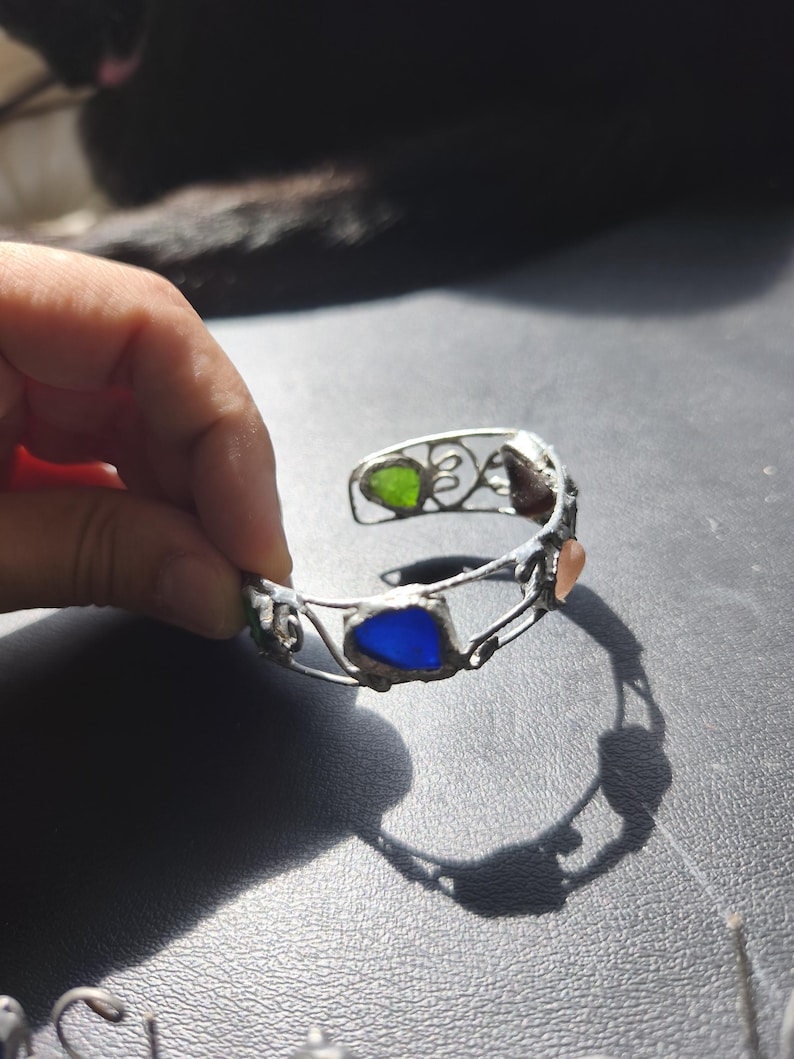 May include: Silver-toned cuff bracelet with colorful glass inserts. The bracelet features green, blue, and peach-colored glass pieces set within a decorative wire frame. The open design allows for easy wear and showcases the vibrant colors of the glass.