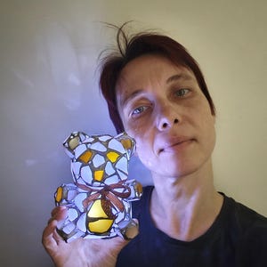 May include: A stained glass teddy bear night light with a brown ribbon. The bear is made of white, yellow, and orange glass pieces. The light is on, illuminating the bear's interior.