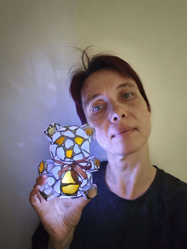 May include: A stained glass teddy bear night light with a brown ribbon. The bear is made of white, yellow, and orange glass pieces. The light is on, illuminating the bear's interior.