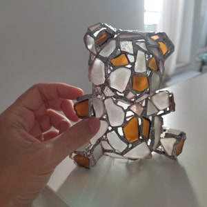 May include: A handcrafted teddy bear sculpture made from pieces of sea glass in white and amber colors, held by a hand. The bear is assembled with a silver-colored metal frame, creating a mosaic effect. The bear is a decorative item.