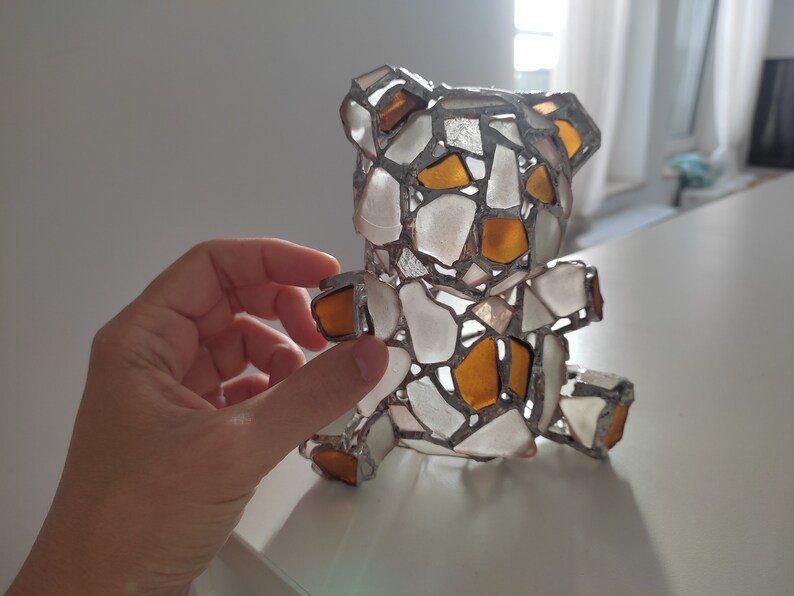 May include: A handcrafted teddy bear sculpture made from pieces of sea glass in white and amber colors, held by a hand. The bear is assembled with a silver-colored metal frame, creating a mosaic effect. The bear is a decorative item.