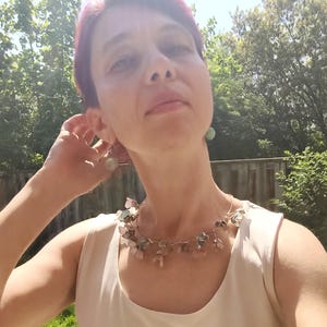 May include: A woman wearing a delicate necklace with small, irregularly shaped stones in shades of pink, green, and clear. The necklace is strung on a thin, silver-colored wire. She is also wearing matching earrings. The woman is wearing a white sleeveless top.