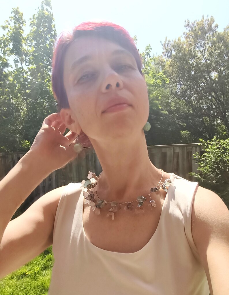 May include: A woman wearing a delicate necklace with small, irregularly shaped stones in shades of pink, green, and clear. The necklace is strung on a thin, silver-colored wire. She is also wearing matching earrings. The woman is wearing a white sleeveless top.