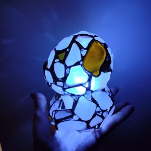 May include: A white and black mosaic lamp with a yellow glass accent. The lamp is lit and glowing blue.