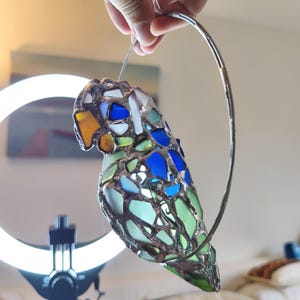 May include: A stained glass parrot ornament with a silver metal ring for hanging. The parrot features a mosaic design with blue, green, white, and yellow glass pieces. The bird's body is primarily green and blue.