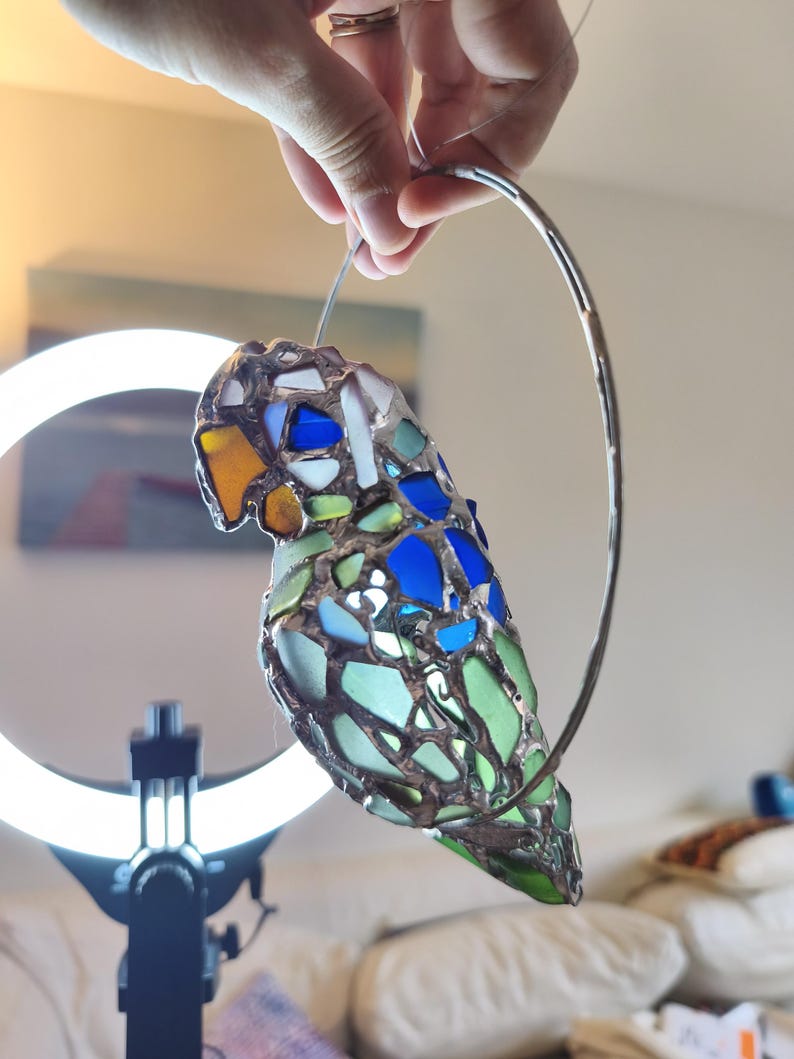 May include: A stained glass parrot ornament with a silver metal ring for hanging. The parrot features a mosaic design with blue, green, white, and yellow glass pieces. The bird's body is primarily green and blue.