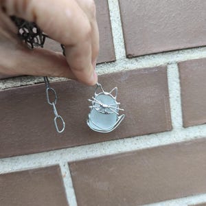 May include: A silver-toned cat pendant necklace. The pendant features a cat silhouette with a light blue, oval-shaped center. The cat's outline and chain are made of silver-colored metal. The necklace is held against a brick wall.
