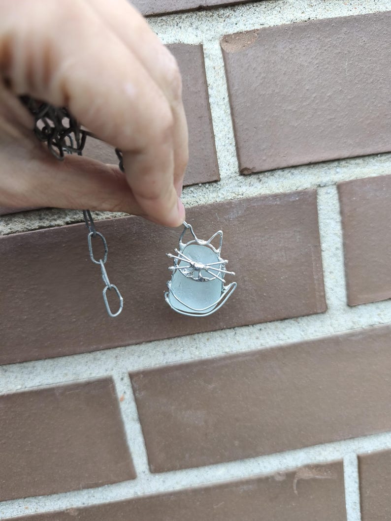May include: A silver-toned cat pendant necklace. The pendant features a cat silhouette with a light blue, oval-shaped center. The cat's outline and chain are made of silver-colored metal. The necklace is held against a brick wall.