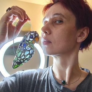 May include: A stained glass parrot ornament with a silver wire hanger. The bird features a mosaic design with blue, green, and orange glass pieces. The parrot is suspended in front of a light.