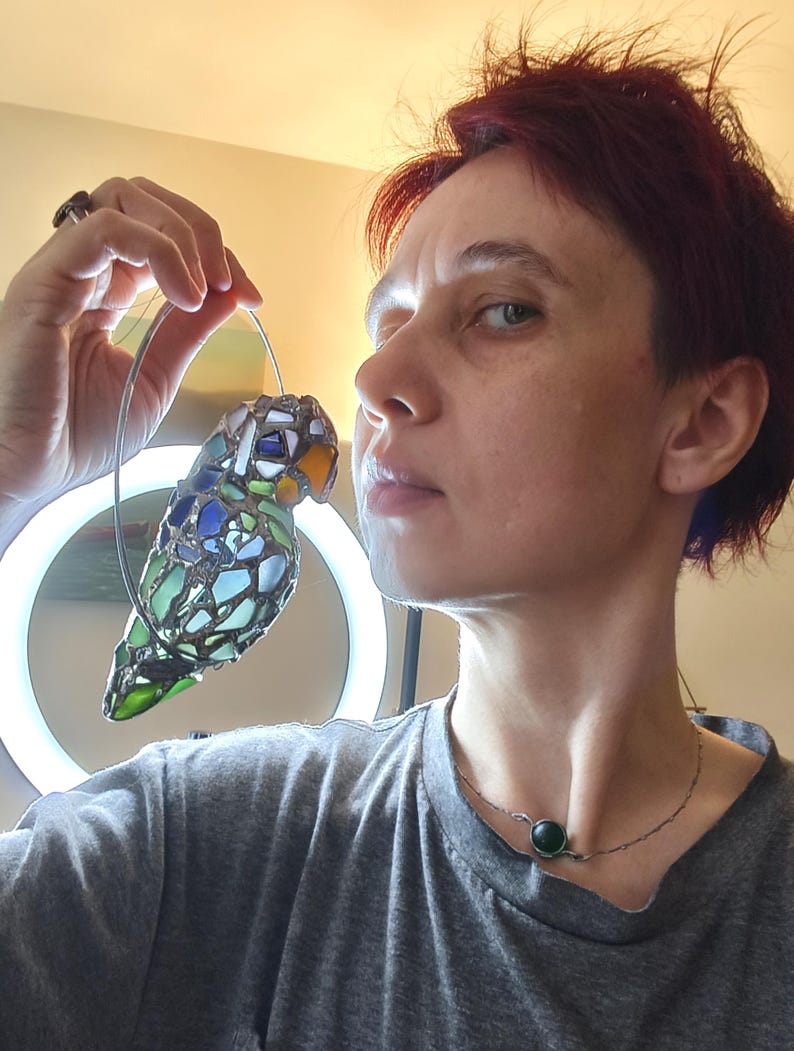 May include: A stained glass parrot ornament with a silver wire hanger. The bird features a mosaic design with blue, green, and orange glass pieces. The parrot is suspended in front of a light.