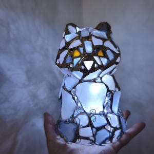 May include: A cat-shaped lamp crafted from white and blue glass pieces, featuring gold accents and yellow eyes. The lamp emits a soft internal glow. The mosaic design is held together by a dark metal frame, creating a unique decorative item.