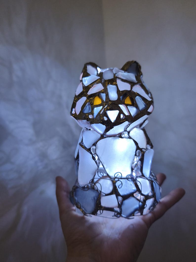 May include: A cat-shaped lamp crafted from white and blue glass pieces, featuring gold accents and yellow eyes. The lamp emits a soft internal glow. The mosaic design is held together by a dark metal frame, creating a unique decorative item.