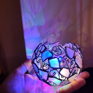 May include: A small, round, glass sphere with a mosaic design. The sphere is made of small, irregular pieces of glass that are held together by a black metal frame. The sphere is lit from within, creating a colorful glow.