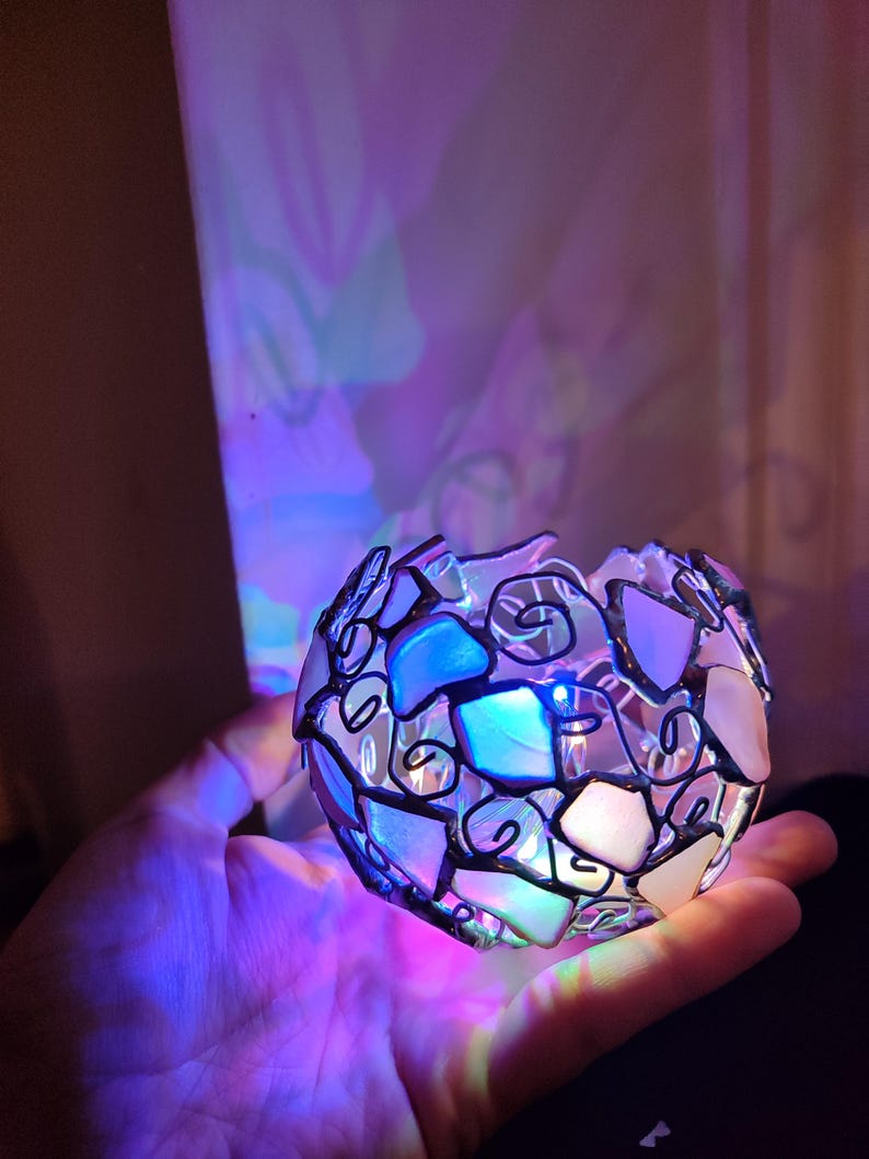 May include: A small, round, glass sphere with a mosaic design. The sphere is made of small, irregular pieces of glass that are held together by a black metal frame. The sphere is lit from within, creating a colorful glow.