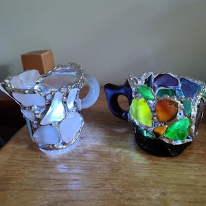 May include: Two small ceramic mugs decorated with colorful mosaic glass pieces. The mug on the left is white with clear glass pieces, while the mug on the right is black with green, yellow, orange, and brown glass pieces.