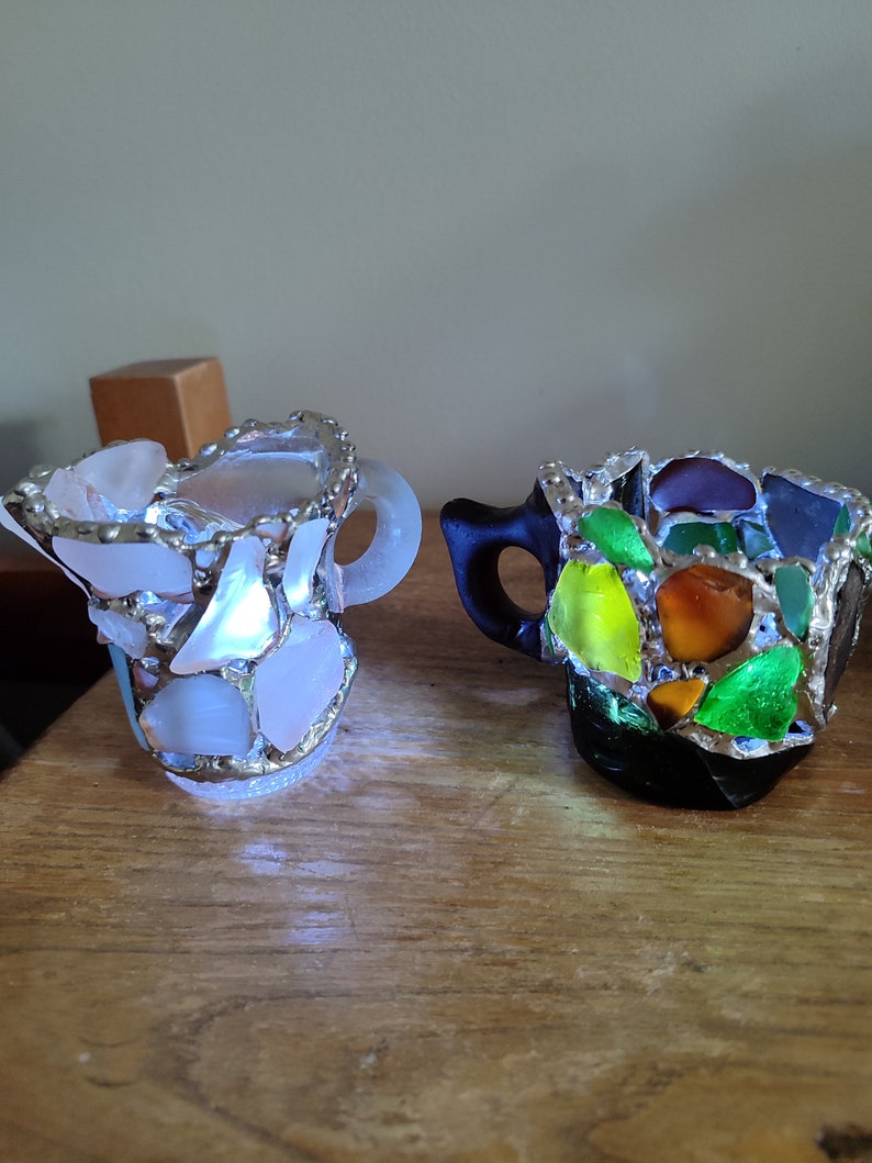 May include: Two small ceramic mugs decorated with colorful mosaic glass pieces. The mug on the left is white with clear glass pieces, while the mug on the right is black with green, yellow, orange, and brown glass pieces.