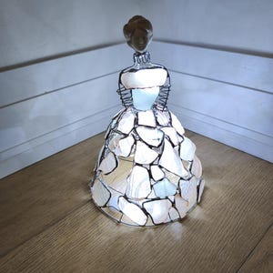 以下が含まれることがあります： 薄茶色の素材で作られた頭部と、暗い金属ワイヤーで繋ぎ合わされた半透明の白いシーグラスのピースで構成されたドレスを持つ人物の装飾フィギュア。内側から光を放つベル型のスカートが特徴です。